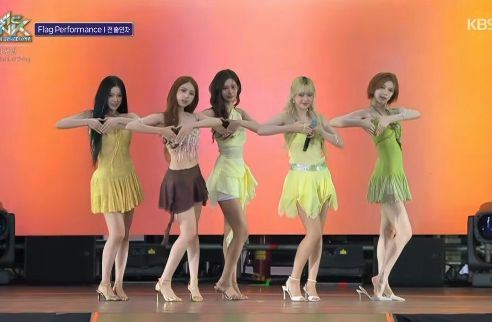 Five female performers in yellow and green outfits make heart shapes with their hands on a stage with an orange backdrop, with "Flag Performance | 전종환자" text and the "KBS MUSIC BANK" logo visible.