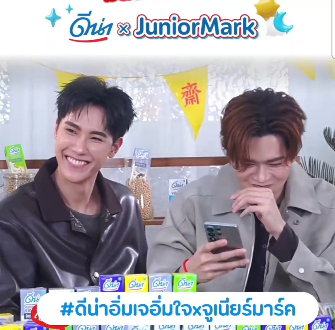 Two young men sit behind a table of colorful snack packs, with the man on the right looking down at a phone while smiling and the man on the left grinning toward the camera. The image appears to be a promotional livestream frame with the Thai logo text “ดีน่า x JuniorMark” across the top and the hashtag “#ดีน่าอิ่มเจอิ่มใจxจูเนียร์มาร์ค” in a large white banner at the bottom, matching the post’s behind-the-scenes caption.