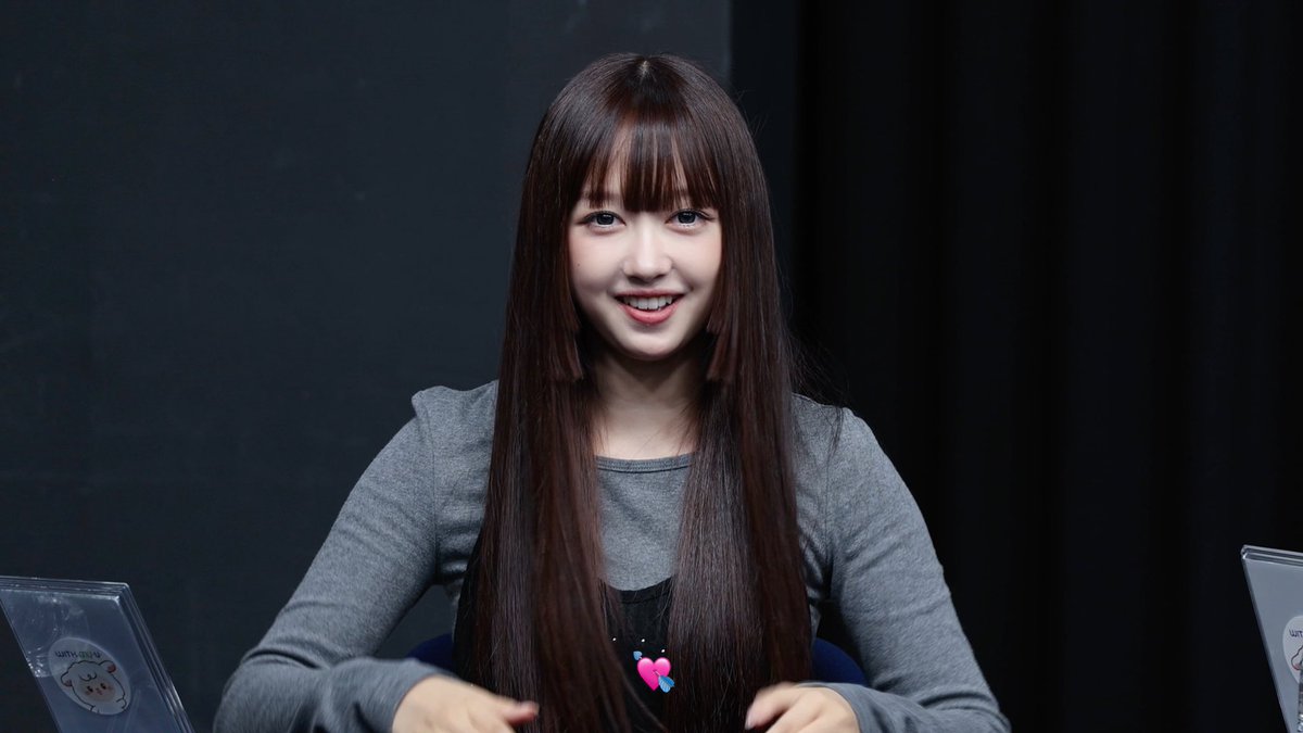 A smiling young woman with long brown hair and bangs, wearing a gray long-sleeve shirt, seated at what appears to be a fan event.