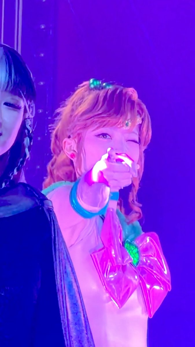 A performer in a Sailor Moon-inspired costume winks and points a glowing glove forward under bright pink and purple stage lighting.