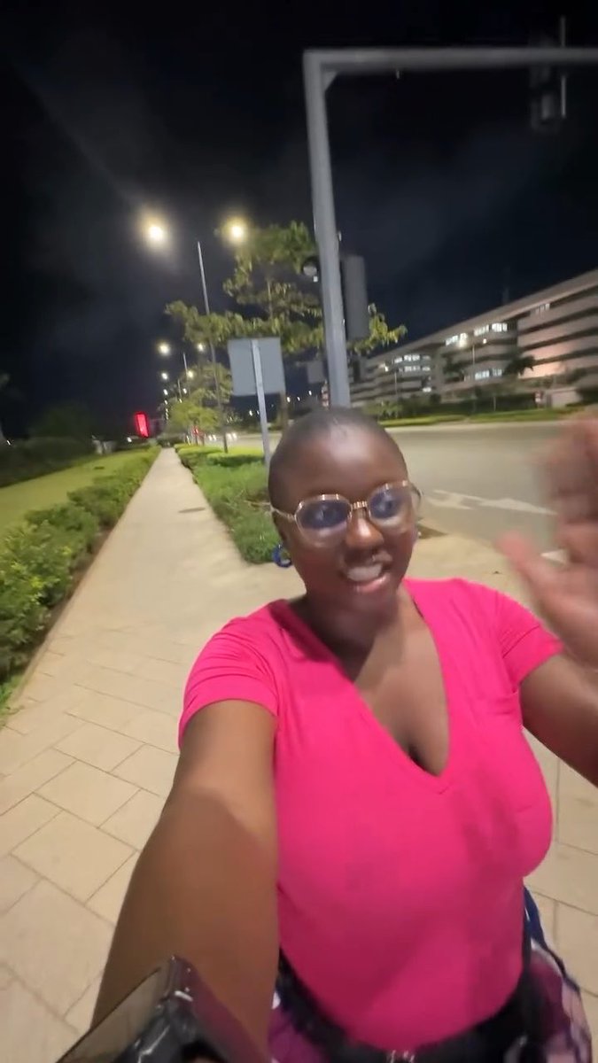 A woman wearing a bright pink v-neck top and glasses takes a selfie while walking on a sidewalk at night, with a city skyline and streetlights in the background.