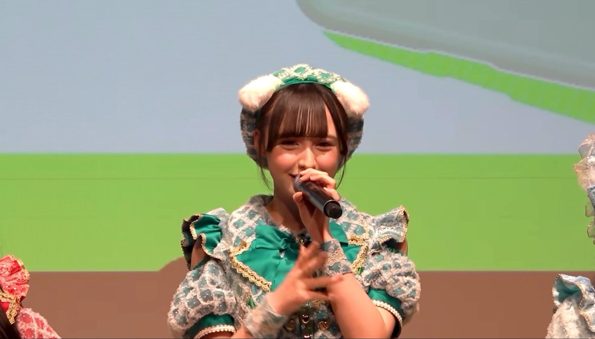 A young woman performs on stage holding a microphone, wearing an ornate green and blue checkered dress with white fur-trimmed headwear against a green and blue background.