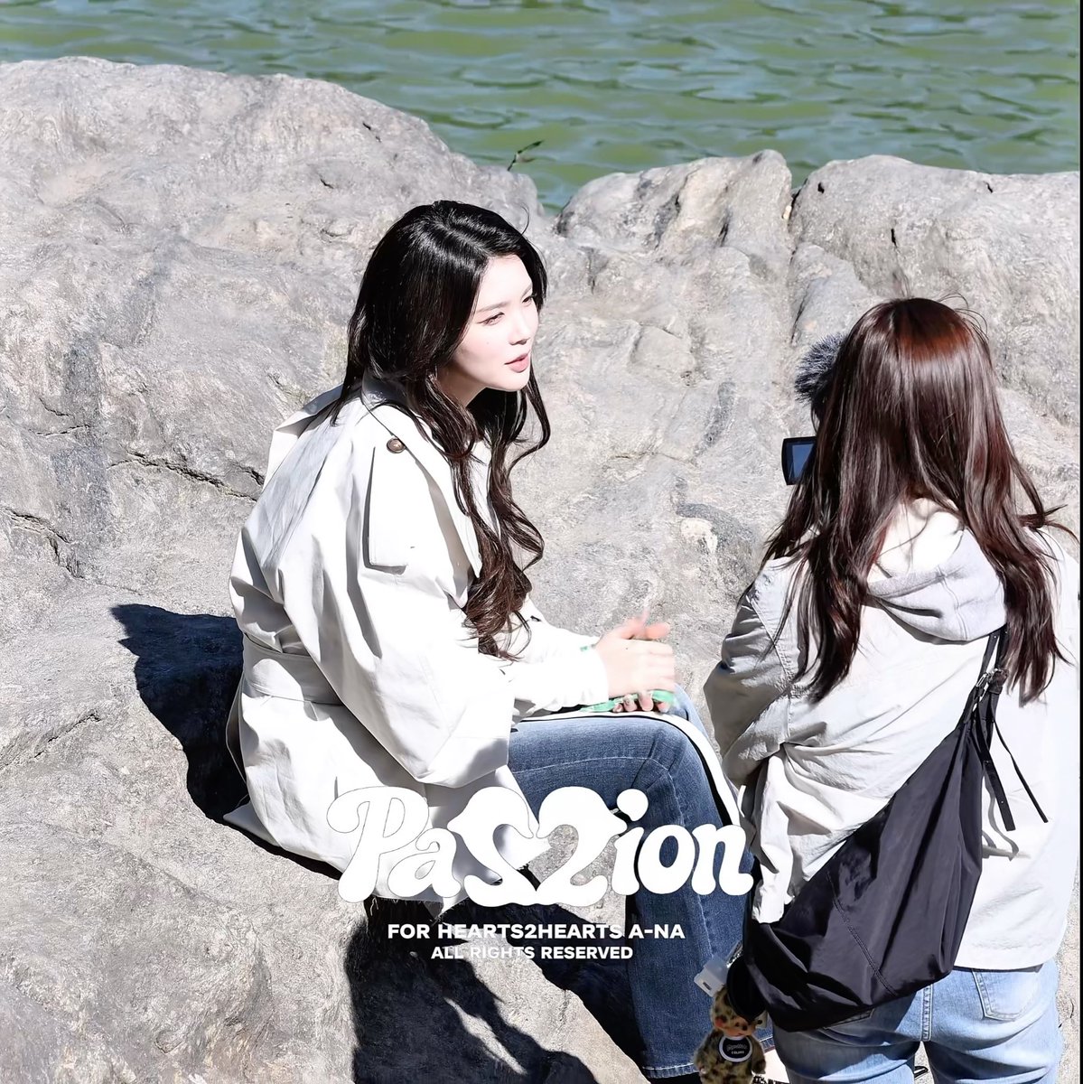 Two women sit on rocks by the water, one in a white trench coat and another with long hair taking her photo, with the image watermarked "PassZion, FOR HEARTS2HEARTS A-NA ALL RIGHTS RESERVED"—aligning with the post tagging Hearts2Hearts member A-NA and Hoon-sung.
