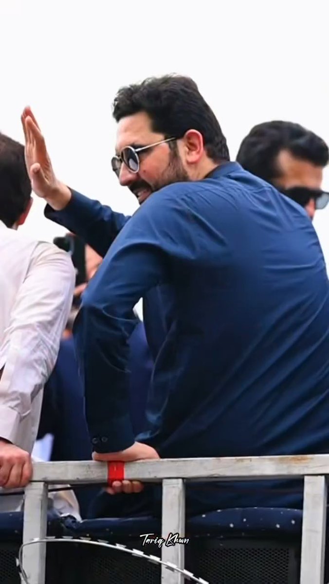 A bearded man wearing sunglasses and a blue shirt waves while leaning against a barrier, with the watermark "TariqKhan" visible at the bottom.
