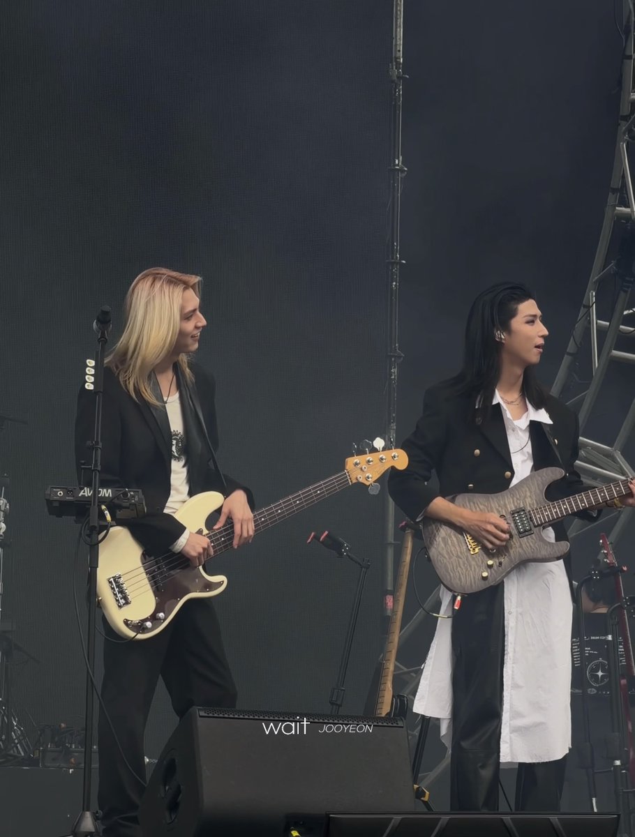Two female musicians perform on stage, one playing a cream bass guitar and the other playing a grey electric guitar, with a watermark reading 'wait JOOYEON' visible.