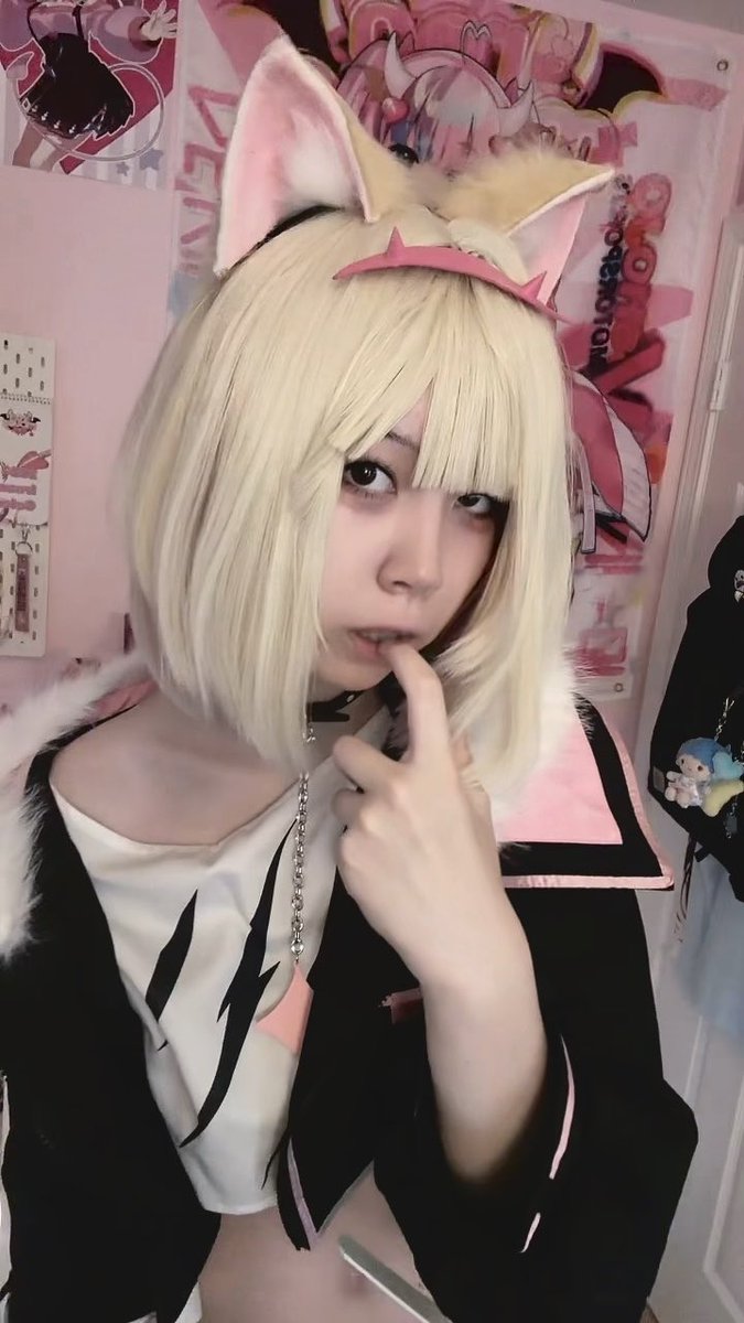 A person with short blonde hair wearing fox ears and a black and white outfit with pink accents, posing with a finger on their lips in front of pink anime-themed decorations and posters.