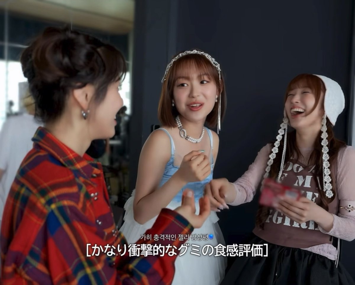 Three young women laugh together in what appears to be a backstage setting, with Japanese subtitles reading '[かなり]衝撃的なグミの食感評価' visible on screen.