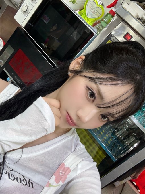 A selfie of Park Jiwon with long black hair and bangs, wearing a white shirt with a floral design, posing in what appears to be a kitchen or shop with a microwave and refrigerators visible in the background.