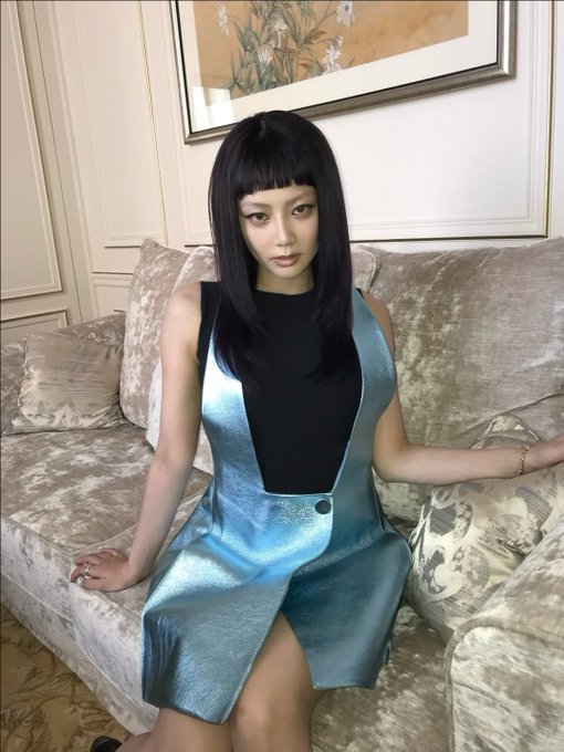 A young woman with long dark hair and bangs sits on a beige couch wearing a shimmering blue and black dress, with a framed artwork visible on the wall behind her.