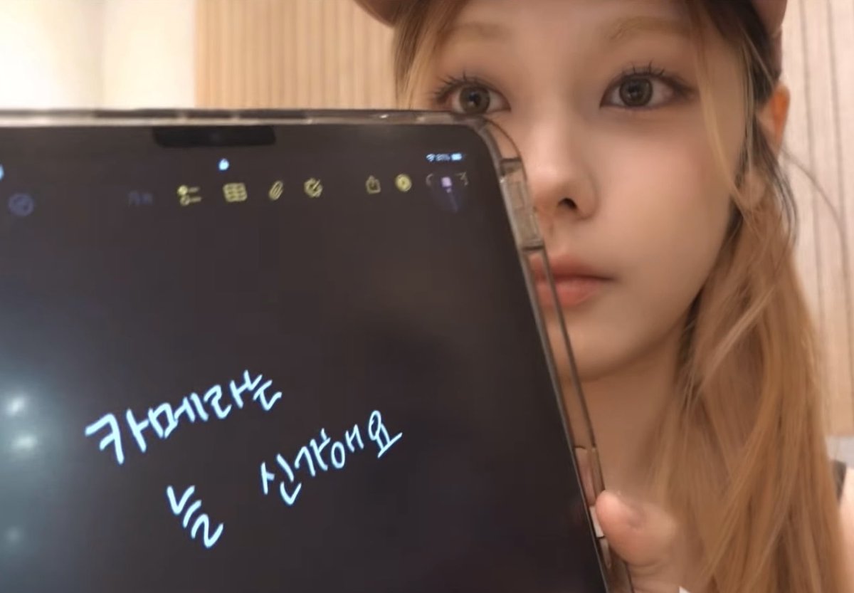 A close-up of a young woman with long blonde hair and large eyes holding up a tablet displaying mirrored handwritten Korean text, including '카메라테스트' and '신기하당요'.