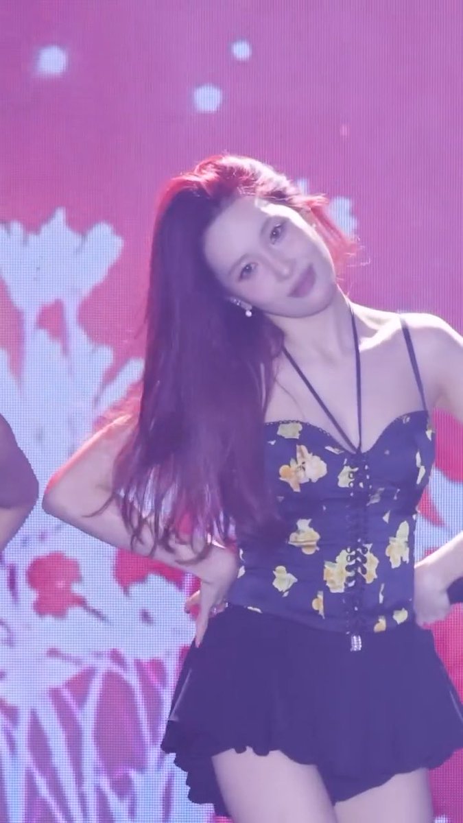 A young woman with long dark hair performs on stage wearing a black floral corset top and black skirt against a vibrant pink and purple lit screen.