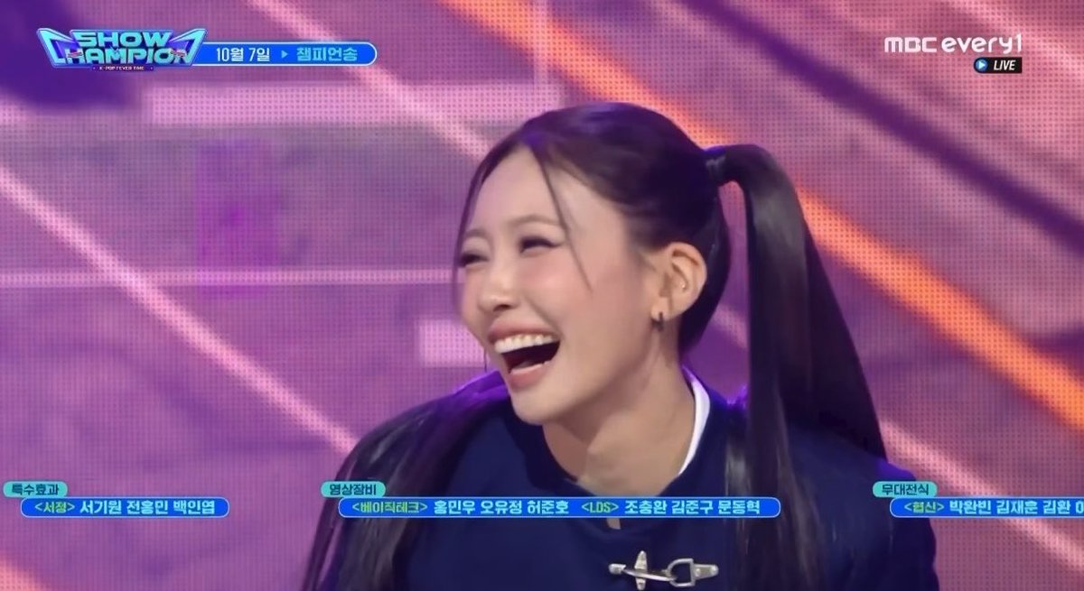 A young woman with pigtails laughs on the Korean music show "Show Champion" broadcast on MBC every1, with Korean text mentioning performances by artists including 권민우 and 조충환.
