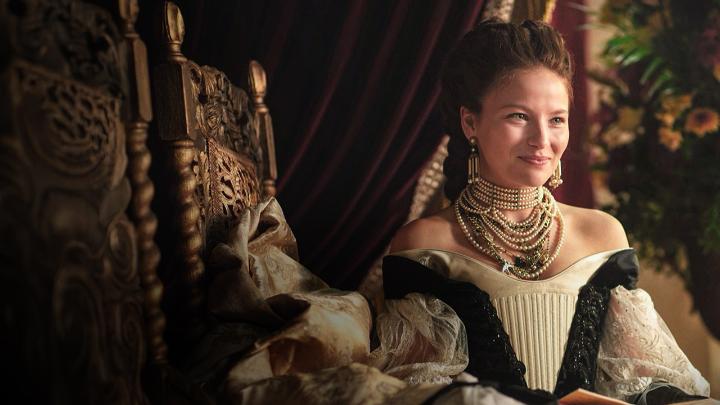 A young woman in an elegant 18th-century gown adorned with pearl necklaces and drop earrings sits regally on an ornately carved wooden chair, smiling softly while holding an open book. The image captures the poised and determined spirit of Empress Elisabeth of Austria, aligning with the post's celebration of her journey in the final season of "The Empress," premiering November 12.