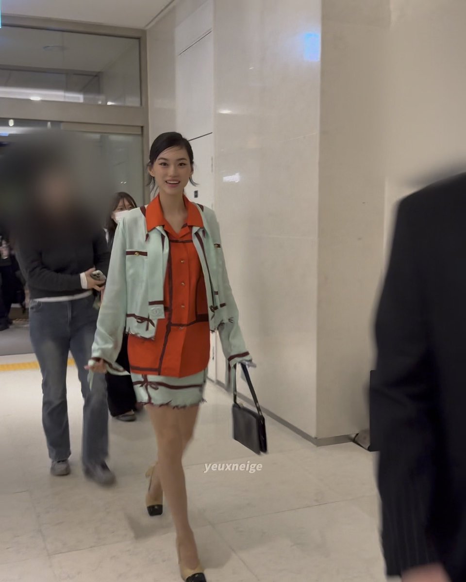 A woman in a mint green and orange tweed outfit smiles while walking through an indoor corridor, carrying a black handbag.