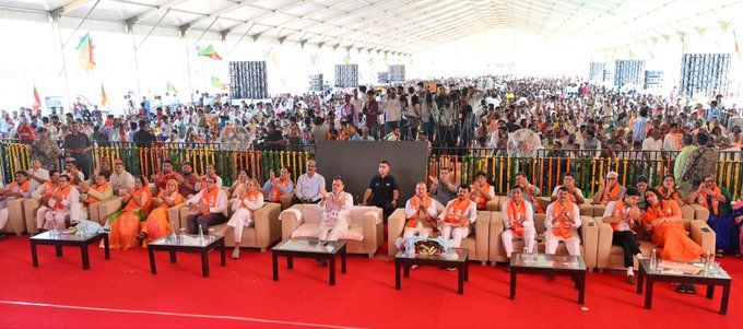 A large crowd gathers under a white tent for what appears to be a religious or cultural event, with seated dignitaries in orange and white robes in the front rows behind a yellow railing.