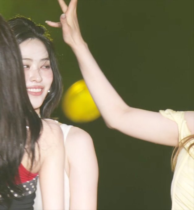 A close-up of a smiling young woman with dark hair on stage, wearing a black top with white polka dots and red accents, with another performer's raised arm visible beside her.