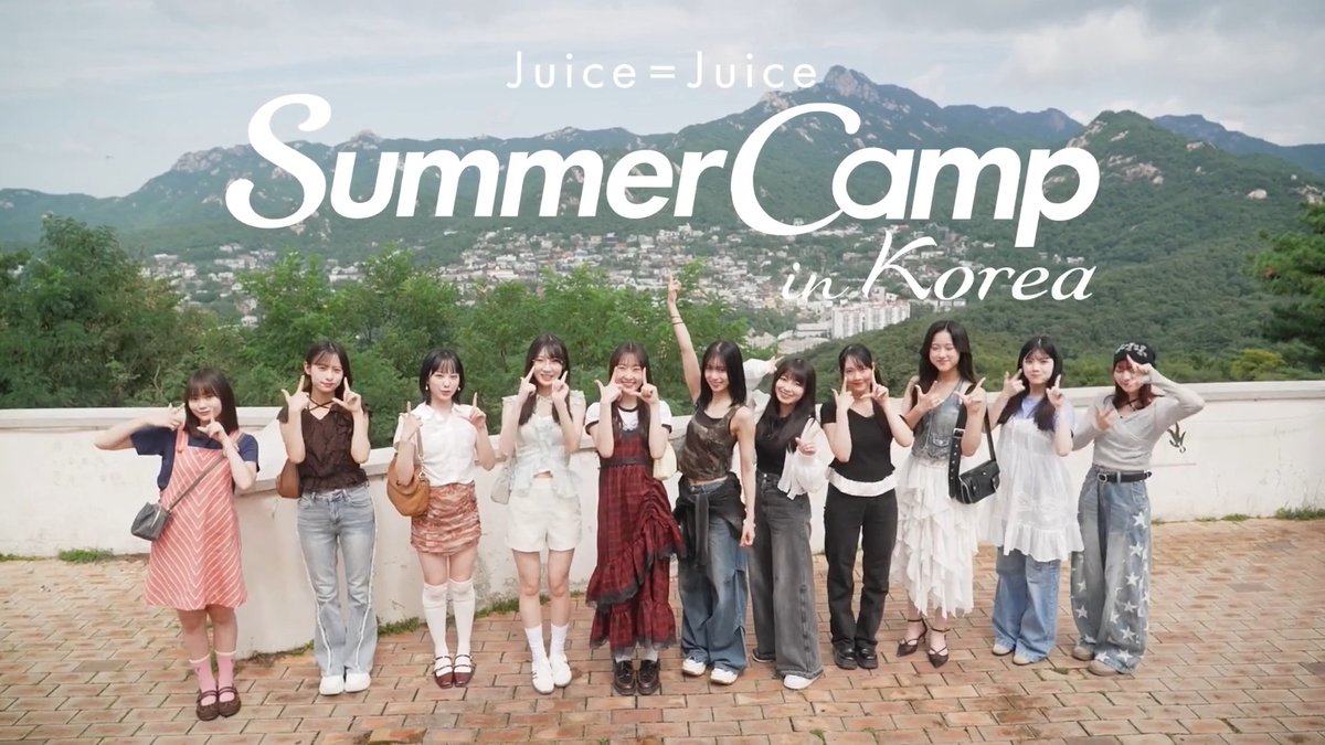 A promotional photo for the J-pop group Juice=Juice titled 'Summer Camp in Korea,' showing eleven members posing outdoors against a mountainous backdrop.