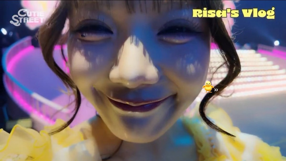 A close-up selfie of a young woman with a playful expression, labeled 'Risa's Vlog' and 'Cutie Street' in the corners, with colorful stage lighting on her face.