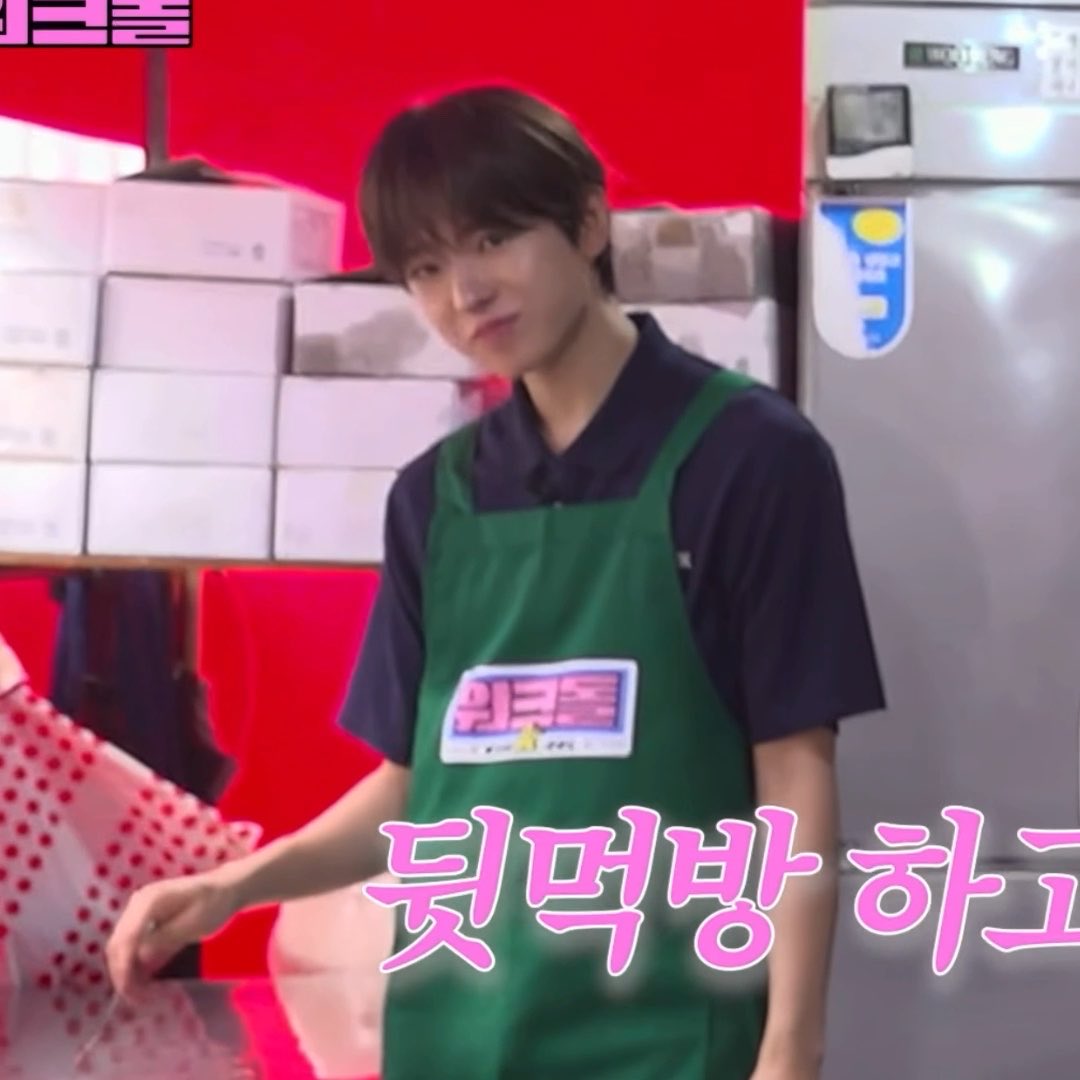 A young man wearing a green apron over a dark polo shirt standing in what appears to be a kitchen or storage area with a refrigerator and stacked boxes; Korean text '뒷먹방 하' is visible.