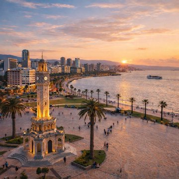 An aerial sunset view of a coastal city featuring a prominent clock tower in a palm-tree-lined plaza along the waterfront.
