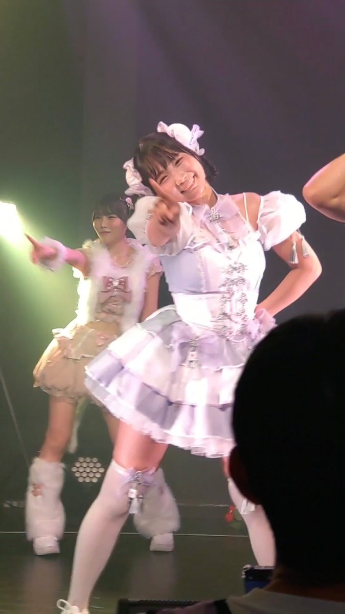 Two performers in elaborate, frilly outfits perform on stage, with the one in the foreground wearing a white and purple dress and smiling brightly while pointing.