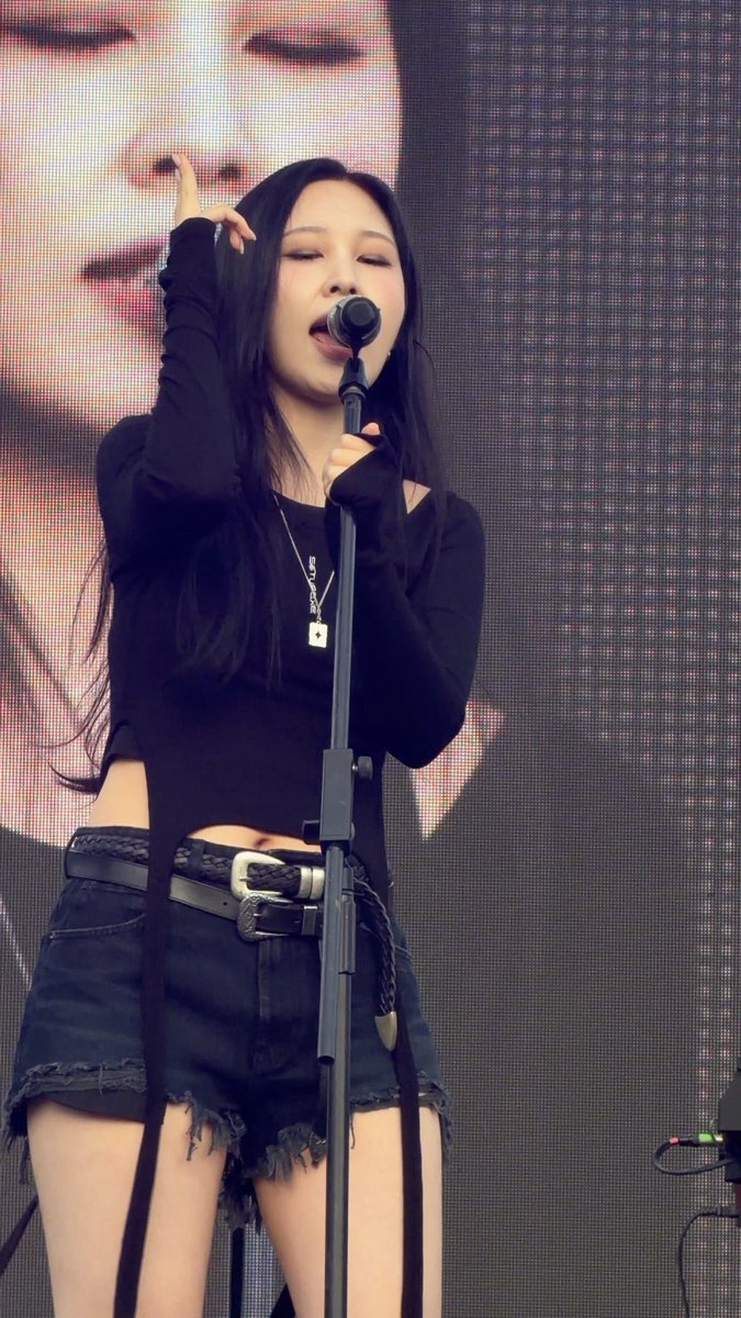 A female K-pop performer sings into a microphone on stage, wearing a black long-sleeve crop top and a distressed denim mini skirt with a large silver buckle belt, against a large LED screen.
