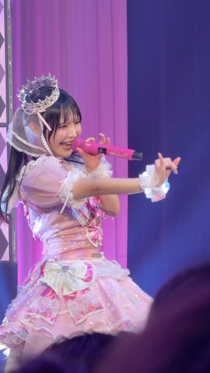 A performer in an elaborate pink and white frilly costume with a tiara sings into a pink microphone on stage against a pink curtain backdrop.
