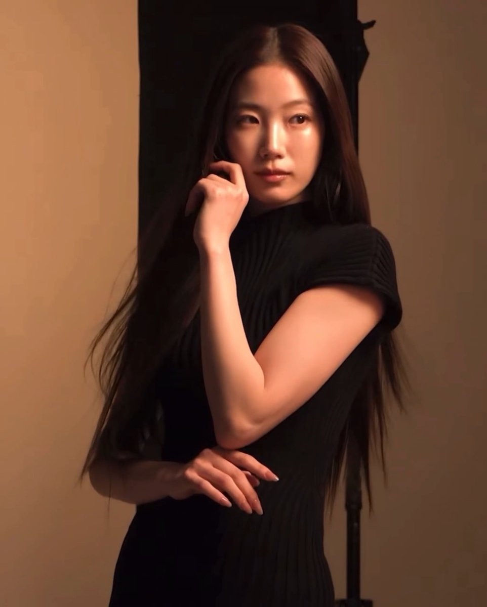 Portrait of a woman with long dark hair wearing a black ribbed top, posed with her hand near her face under warm lighting.