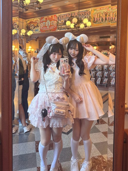 Two young women in matching pastel frilly dresses with white bunny ear headbands pose together for a mirror selfie in what appears to be a vintage-style venue with ornate chandeliers and a checkered floor, with the hashtag referencing "Sayaka Tanizaki Birthday Festival 2026."