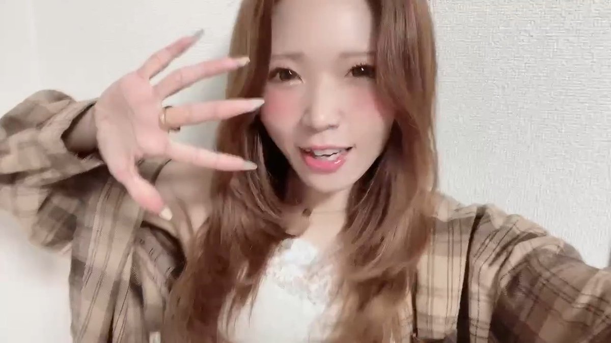 A woman with long brown hair wearing a plaid overshirt holds up her hand with long manicured nails toward the camera.