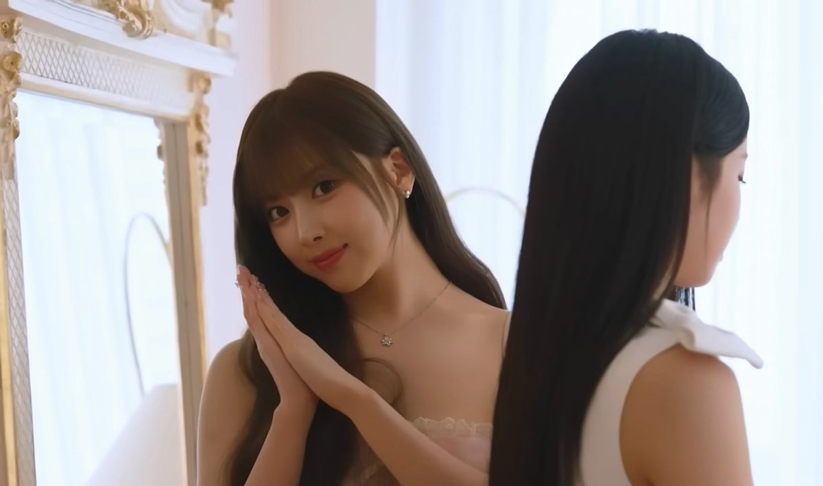 A young woman with long brown hair wearing a light-colored top smiles softly while posing in front of an ornate mirror, with another person visible in the foreground.