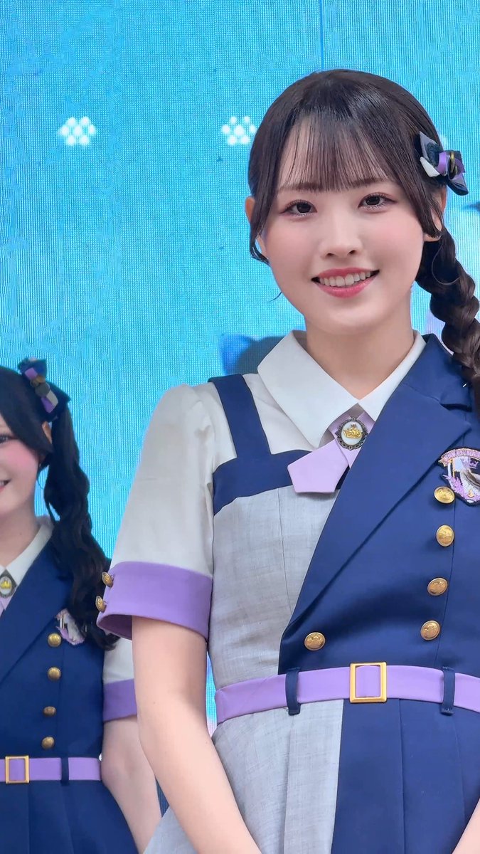 A young woman in a blue and white uniform with gold buttons and purple accents smiles at the camera, with another person in matching attire partially visible to her left.