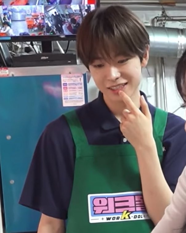 The same young man from image 9, now wearing a green apron with Korean text '워크돌' (Workdol) over his dark shirt, appearing on what looks like a variety show set with a playful expression.