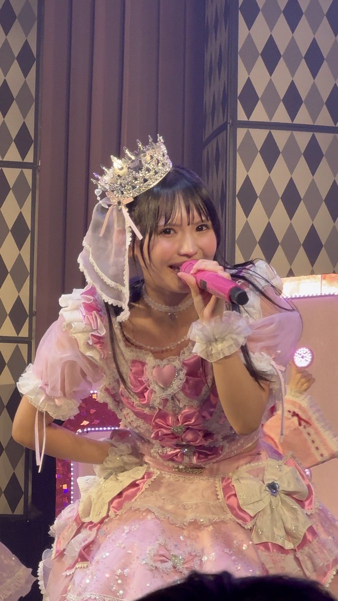 A performer in an elaborate pink frilly dress with a crystal tiara and matching headpiece sings into a pink microphone on a stage with diamond-patterned walls.