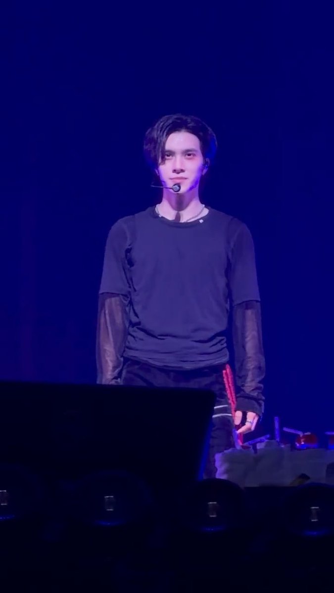 A performer on stage wearing a headset microphone and a layered dark top with sheer sleeves appears to be smiling slightly, matching the caption about Hendery trying to contain his expression while Xiaojun unbuttons his clothes.