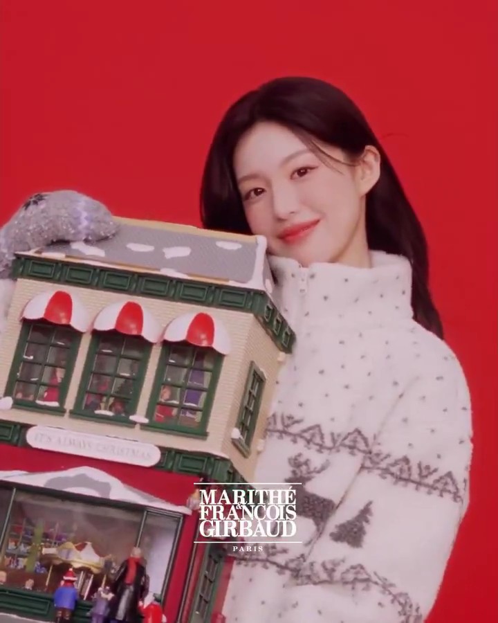 A promotional image for "MARITHÉ FRANÇOIS GIRBAUD PARIS" showing a woman in a white textured jacket holding a miniature Christmas village house against a bright red background.