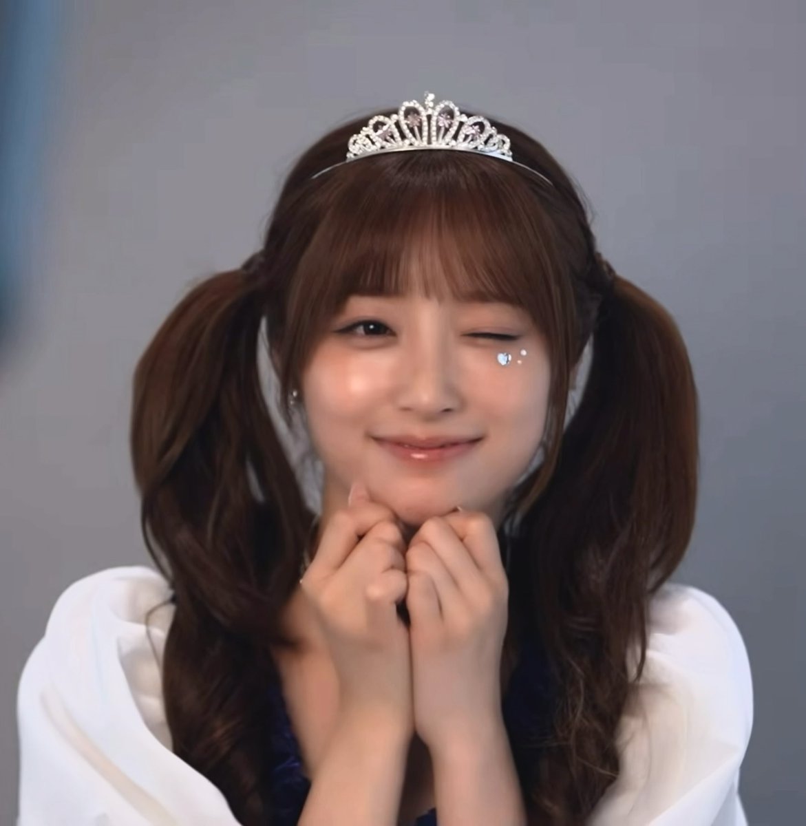 A woman wearing a tiara with her hands near her face winks and smiles, featuring a decorative gem under her closed eye.