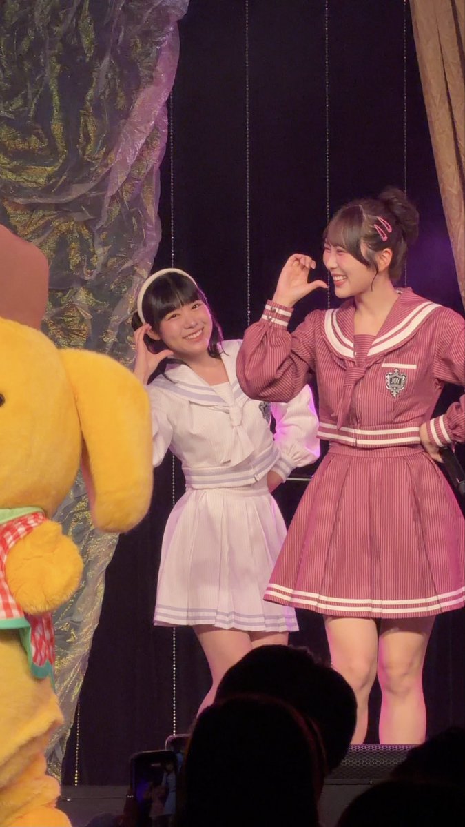 Two young women in sailor-style outfits—one in white, one in pink—pose playfully on stage with a yellow mascot character nearby.
