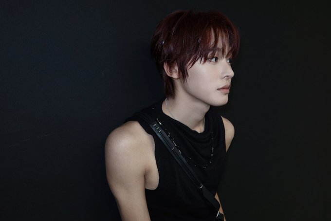 A portrait of a young man with reddish-brown hair wearing a black sleeveless top with straps against a black background.