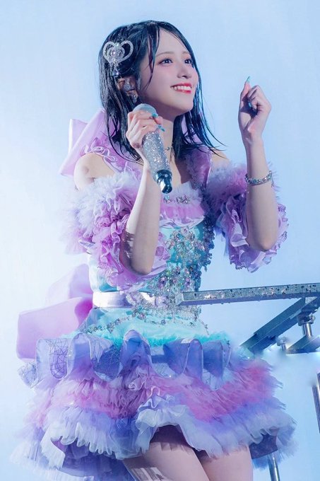 A young female performer in an elaborate pastel purple, blue, and pink ruffled dress smiles while holding a silver microphone on stage, wearing a sparkly hair accessory.