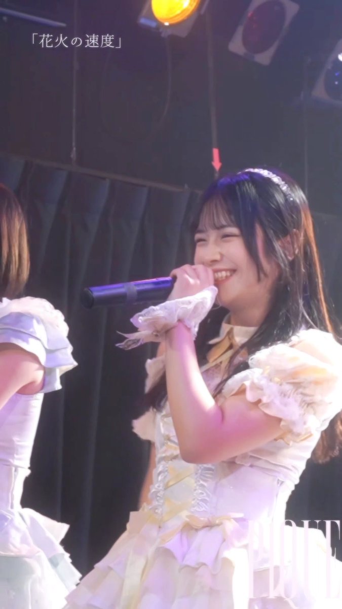 A performer with long dark hair sings into a microphone on stage wearing a white frilly outfit, with Japanese text '「花火の速度」' displayed in the upper left corner.