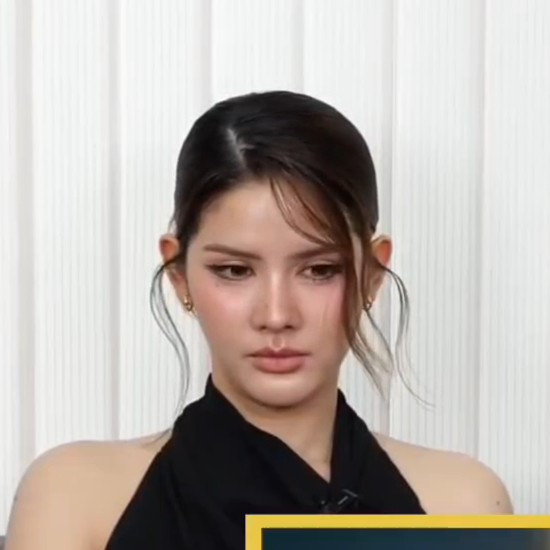 A close-up of a young woman with dark hair pulled back, wearing a black halter top, looking slightly downward against a white ribbed background.