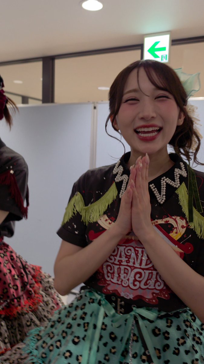 A smiling young woman in a teal leopard-print dress and fringed top presses her hands together, with another similarly dressed person partially visible to her left.
