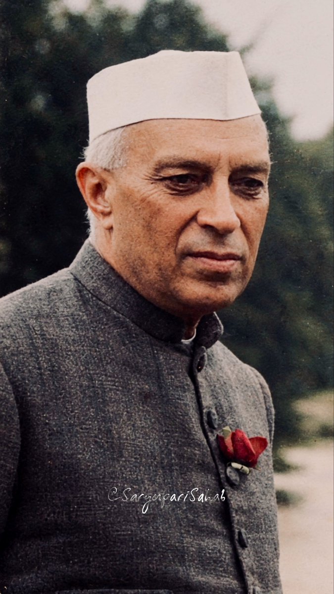 A colorized historical portrait of an elderly man wearing a white Gandhi cap and a grey buttoned jacket with a red flower pinned to the lapel, watermarked with 'SangeetvariSainii'.