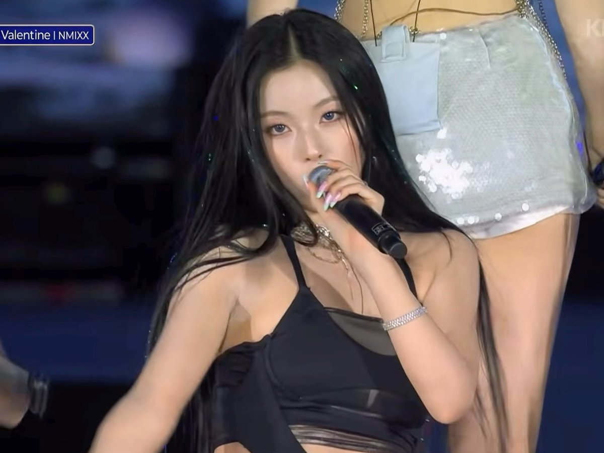 A K-pop performer sings into a microphone on stage, labeled 'Valentine | NMIXX' in the corner.