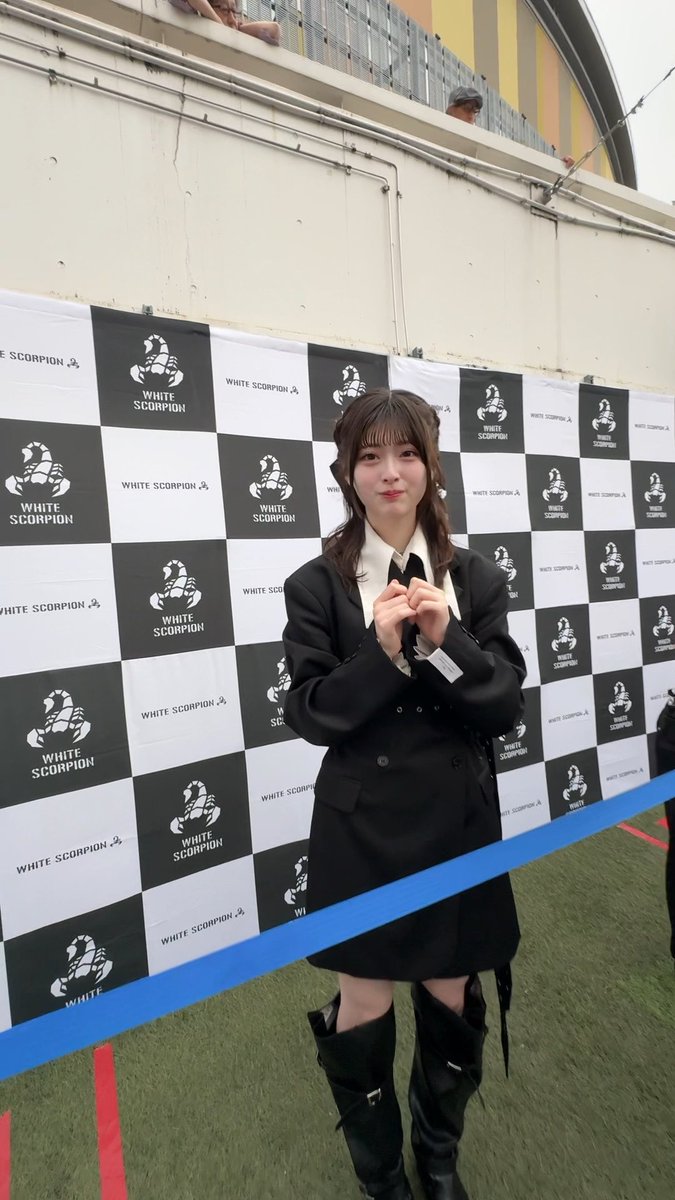 A young woman in a black blazer, skirt, and thigh-high boots poses in front of a checkered backdrop displaying the "WHITE SCORPION" logo repeatedly.
