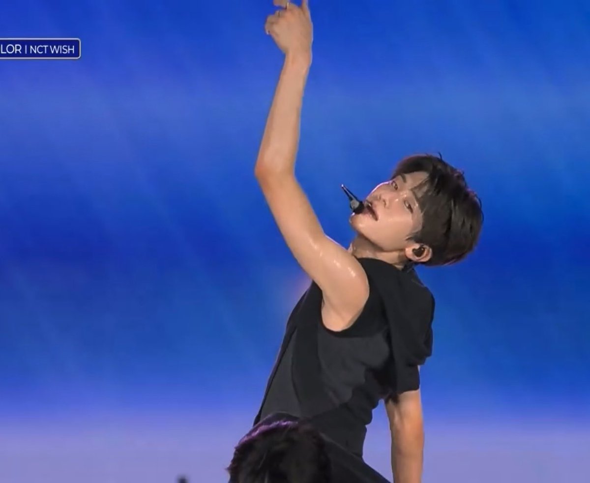 A performer in a black sleeveless top strikes a dramatic pose with one arm raised overhead on stage against a blue backdrop, with "NCT WISH" visible in the top-left watermark. The image captures an intense moment of the choreography, likely from SION's solo part referenced in the caption.