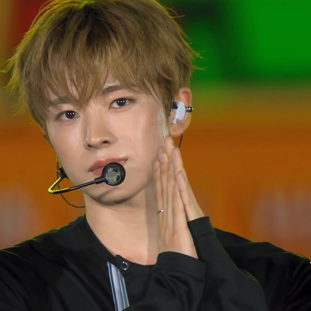 A close-up shot of a male K-pop idol with light brown hair wearing a headset microphone and an in-ear monitor, posing with his hands together during a performance.