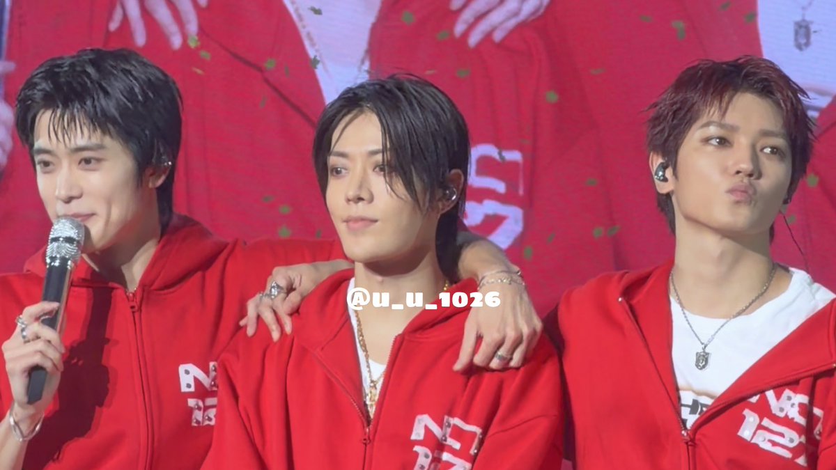Three young men in red hoodies performing on stage, with the one on the left holding a microphone and a watermark reading '@u_u_1026'.