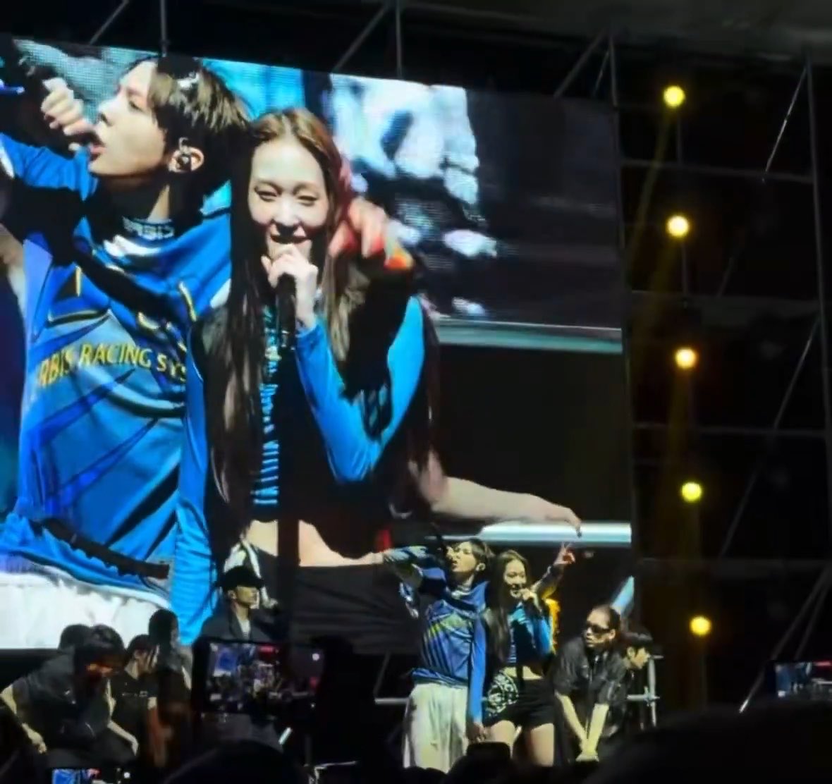 A concert performance is shown on a large screen featuring two performers, one male in a blue jersey and one female with long hair holding a microphone, while below them on stage other performers and crew members are visible.